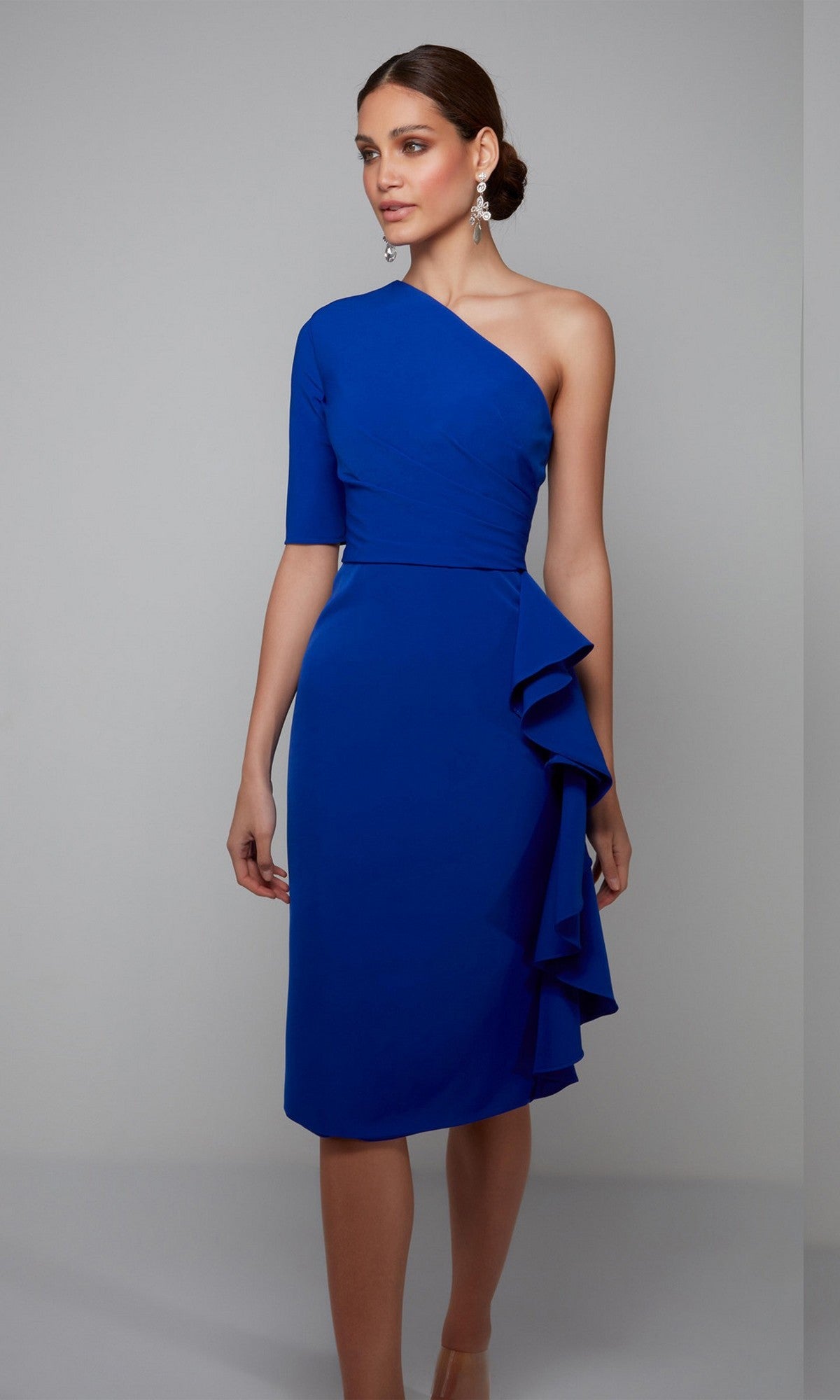 A woman stands against a plain gray background wearing the Alyce 70006 knee-length, semi-formal dress in royal blue with a one-shoulder design and ruffled skirt detail.